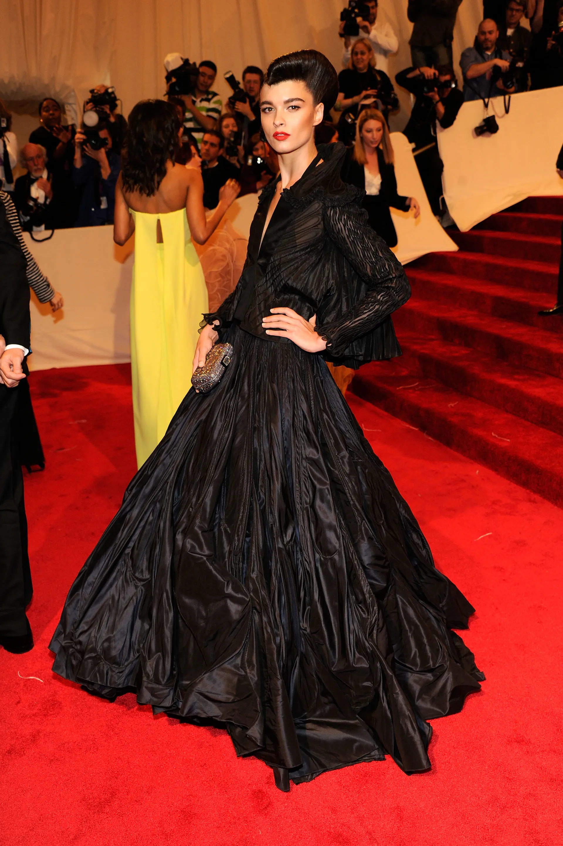 red carpet photo of slender white woman, wearing a volumous black gown with long sleeves and deep v-neck featuring interesting pleated details, glittery silver clutch, dark hair swept up into an odd sort of roll or wave, clean eyes, strong brows, clean orange-red lip
